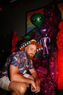 A man in a Hawaiin shirt crouched down posing in front of a temporary sculpture with two inflatable alien figures on it
