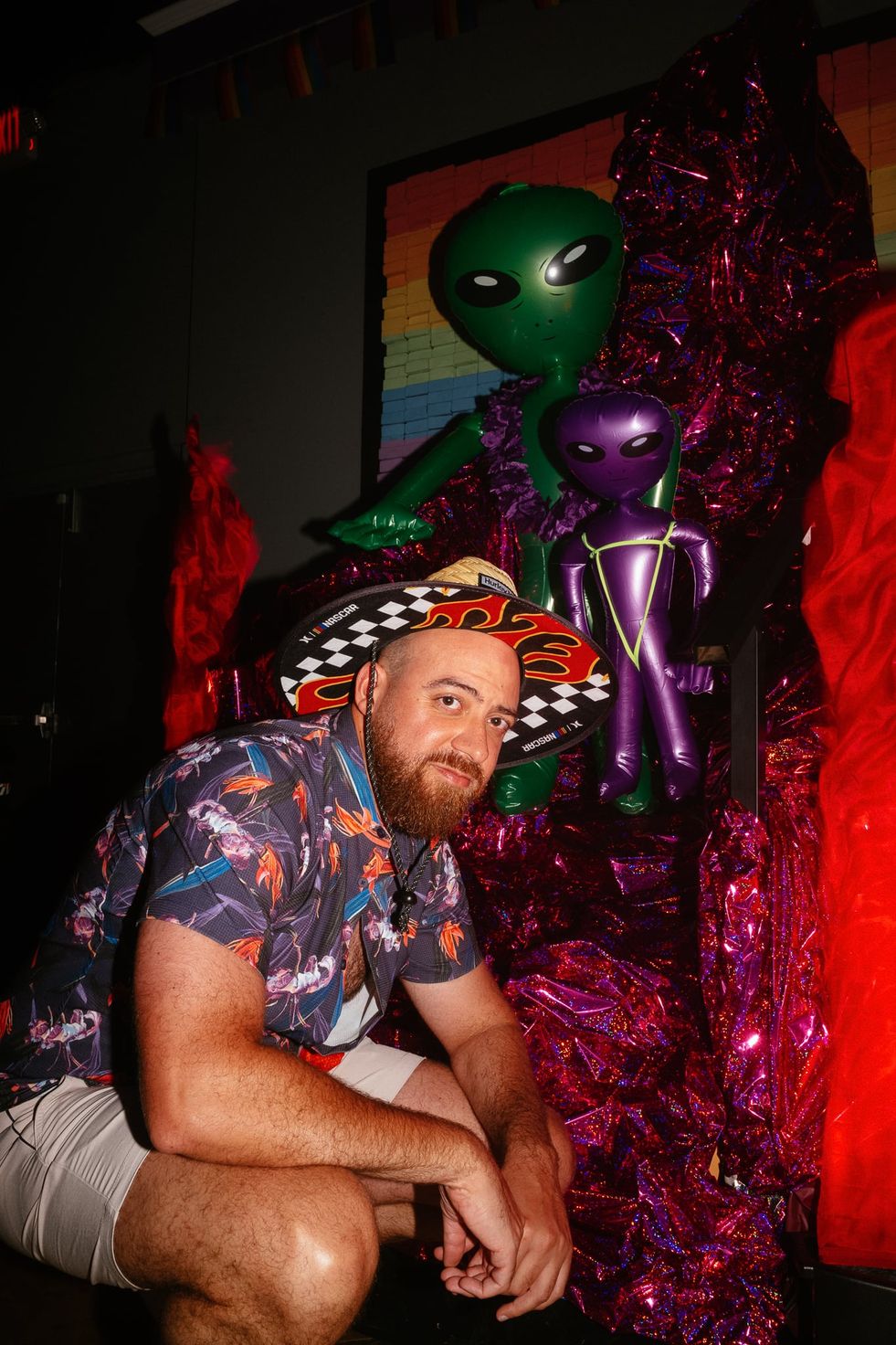 A man in a Hawaiin shirt crouched down posing in front of a temporary sculpture with two inflatable alien figures on it