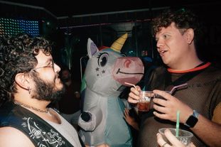 Two men in a club having a conversation, another person in the background is wearing an inflatable unicorn costume