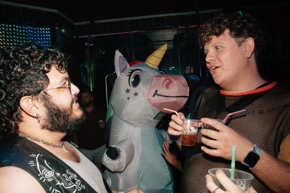 Two men in a club having a conversation, another person in the background is wearing an inflatable unicorn costume