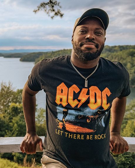 A man wearing an AC/DC shirt, leaning on an overlook's railing. A broad river is in the background.