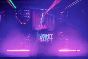 A DJ in the booth in a club. The screen below displaying the Night Shift logo
