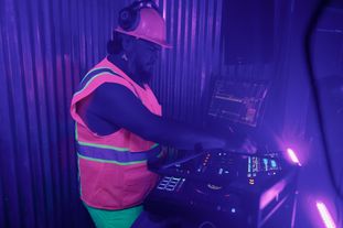 A DJ performing wearing a glowing workwear vest and hardhat, with small penis earrings