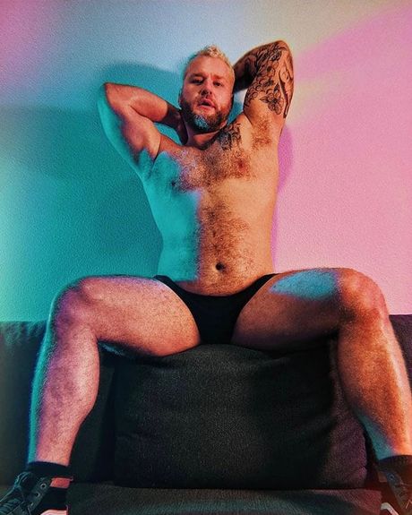 A tattooed man with a beard sitting down with his arms up in a sexy pose