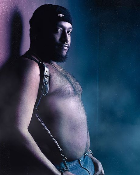 A shirtless man in jeans and a harness leaning against a wall.