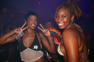 Two women posint for the camera, the woman on the left is holding up peace sines and her diamond studded tooth is shimmering from the camera flash
