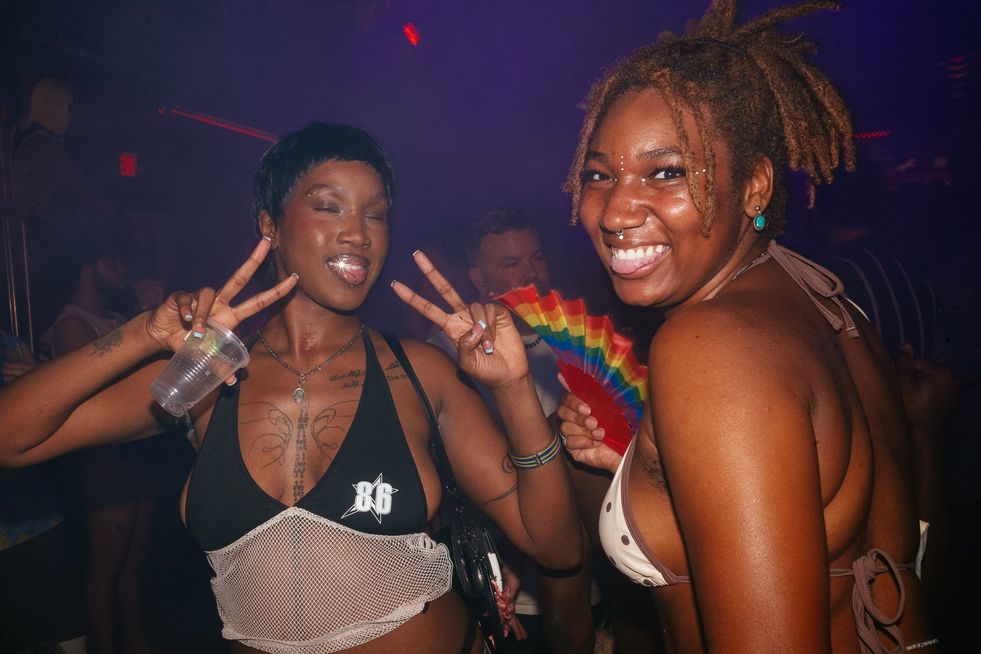 Two women on the dance floor. One making peace signs with her hands, her diamond tooth makes a star shape from the flash