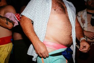 A thick hairy man on the dance floor with his shirt unbuttoned, pulling his shorts down to reveal a pink Speedo
