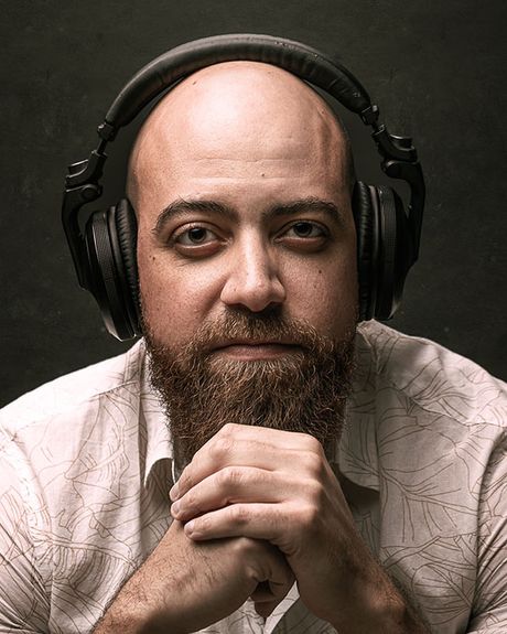 A bearded man wearing headphones with his hands cupped at his chin.