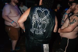 A group of men dancing in a club. One has a leather vest with a Keith Haring style painting on the back