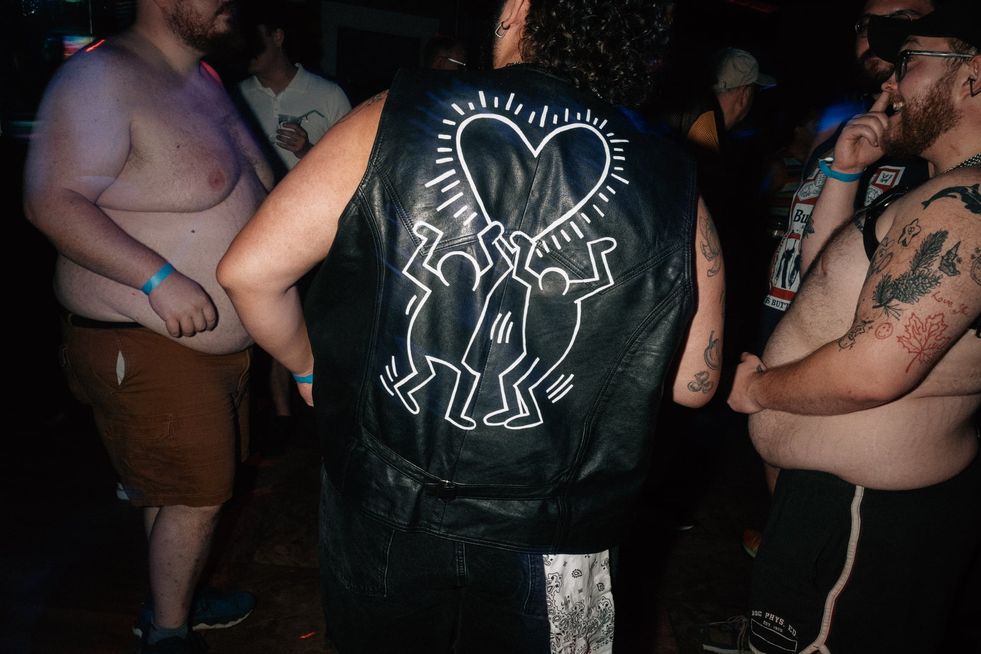 A group of men dancing in a club. One has a leather vest with a Keith Haring style painting on the back